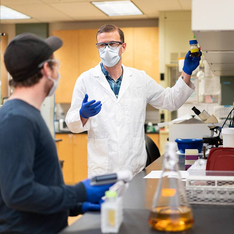A professor wearing lab gear shows a beaker of liquid to a student.